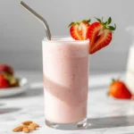A tall glass of creamy strawberry almond milk garnished with fresh strawberry slices on a white marble countertop